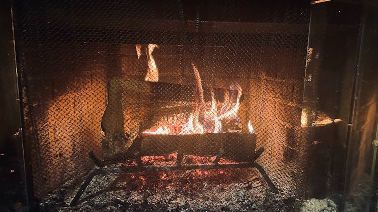 A warm and cozy fire burns brightly within a fireplace, surrounded by a protective mesh screen. The flames cast a gentle glow, illuminating the brick interior of the fireplace. Logs are arranged on a metal grate, contributing to the flickering firelight and emitting a comforting ambiance.