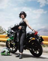 A person wearing black clothing stands confidently beside a green and black motorcycle on a road. The gleaming motorcycle has detailed decals, and a matching helmet is placed on the ground. The sky is bright with scattered clouds, accentuating the edgy, rebellious vibe.