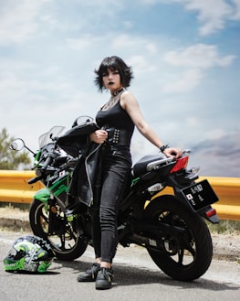 A person wearing black clothing stands confidently beside a green and black motorcycle on a road. The gleaming motorcycle has detailed decals, and a matching helmet is placed on the ground. The sky is bright with scattered clouds, accentuating the edgy, rebellious vibe.