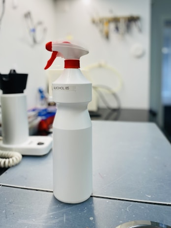 A white spray bottle labeled 'ALKOHOL 85' is placed on a grayish work surface. The bottle has a red and white nozzle. In the background, there are various indistinct objects including another bottle, a coiled cable, and what appears to be a wall-mounted organizer with tools.