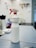 A white spray bottle labeled 'ALKOHOL 85' is placed on a grayish work surface. The bottle has a red and white nozzle. In the background, there are various indistinct objects including another bottle, a coiled cable, and what appears to be a wall-mounted organizer with tools.