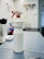 A white spray bottle labeled 'ALKOHOL 85' is placed on a grayish work surface. The bottle has a red and white nozzle. In the background, there are various indistinct objects including another bottle, a coiled cable, and what appears to be a wall-mounted organizer with tools.