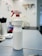 A white spray bottle labeled 'ALKOHOL 85' is placed on a grayish work surface. The bottle has a red and white nozzle. In the background, there are various indistinct objects including another bottle, a coiled cable, and what appears to be a wall-mounted organizer with tools.