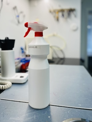 A white spray bottle labeled 'ALKOHOL 85' is placed on a grayish work surface. The bottle has a red and white nozzle. In the background, there are various indistinct objects including another bottle, a coiled cable, and what appears to be a wall-mounted organizer with tools.