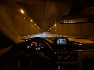 View from the driver's seat of a car traveling through a tunnel, with illuminated dashboard displaying various controls and a GPS screen. The road is dimly lit by overhead lights and another car is visible in the lane ahead.
