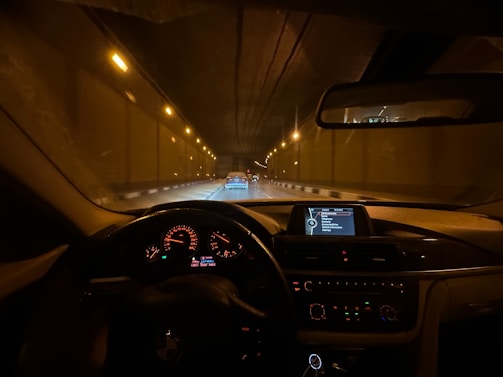 View from the driver's seat of a car traveling through a tunnel, with illuminated dashboard displaying various controls and a GPS screen. The road is dimly lit by overhead lights and another car is visible in the lane ahead.