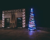 A nighttime scene featuring a brightly lit, decorated Christmas tree next to a building with the name 'Huanchaco' displayed on it. The tree is adorned with multicolored lights and a star at the top. The building has wooden architectural elements and is decorated with circular patterns featuring stylized faces.