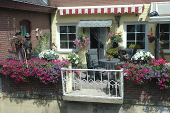 A charming balcony with a table and two chairs decorated with colorful flowers. The railing is adorned with vibrant pink, purple, and white flowers. A small statue of a girl sits on one of the chairs, adding a quaint touch. The building exterior features a red and beige striped awning and pale yellow walls, with decorative wrought iron details.