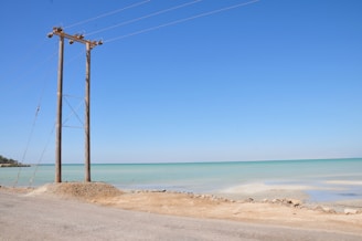 A coastal scene featuring a tall wooden utility pole with multiple power lines extending into the distance. The ocean is visibly calm with a light turquoise hue, meeting a clear blue sky above. The sandy shore and a rough road run alongside the water, with sparse vegetation on the far left.