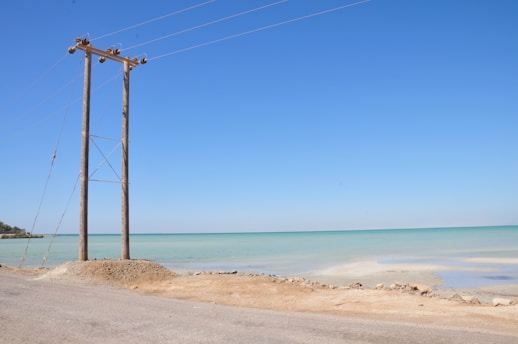 A coastal scene featuring a tall wooden utility pole with multiple power lines extending into the distance. The ocean is visibly calm with a light turquoise hue, meeting a clear blue sky above. The sandy shore and a rough road run alongside the water, with sparse vegetation on the far left.