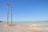 A coastal scene featuring a tall wooden utility pole with multiple power lines extending into the distance. The ocean is visibly calm with a light turquoise hue, meeting a clear blue sky above. The sandy shore and a rough road run alongside the water, with sparse vegetation on the far left.