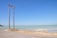 A coastal scene featuring a tall wooden utility pole with multiple power lines extending into the distance. The ocean is visibly calm with a light turquoise hue, meeting a clear blue sky above. The sandy shore and a rough road run alongside the water, with sparse vegetation on the far left.