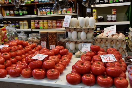 A market stall displays a variety of produce and goods. Fresh red tomatoes are neatly arranged at the forefront, with handwritten price signs in a foreign language placed on top. Behind the tomatoes, stacks of white and brown eggs are visible, with their own signs. Shelves in the background are filled with jars and packaged goods in multiple colors, adding a vibrant backdrop to the scene. Glass jars filled with nuts and dried fruits are positioned between the produce, indicating a diverse offering.