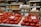 A market stall displays a variety of produce and goods. Fresh red tomatoes are neatly arranged at the forefront, with handwritten price signs in a foreign language placed on top. Behind the tomatoes, stacks of white and brown eggs are visible, with their own signs. Shelves in the background are filled with jars and packaged goods in multiple colors, adding a vibrant backdrop to the scene. Glass jars filled with nuts and dried fruits are positioned between the produce, indicating a diverse offering.