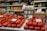 A market stall displays a variety of produce and goods. Fresh red tomatoes are neatly arranged at the forefront, with handwritten price signs in a foreign language placed on top. Behind the tomatoes, stacks of white and brown eggs are visible, with their own signs. Shelves in the background are filled with jars and packaged goods in multiple colors, adding a vibrant backdrop to the scene. Glass jars filled with nuts and dried fruits are positioned between the produce, indicating a diverse offering.