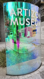 A vibrant, iridescent glass panel with the words 'Artizon Museum' is installed in an urban setting. The surrounding environment reflects off the glass, creating a colorful blend of blues, greens, and purples. The ground is paved with large gray tiles, and there are patches of greenery nearby. The background hints at a busy cityscape with buildings and vehicles visible through the reflections.