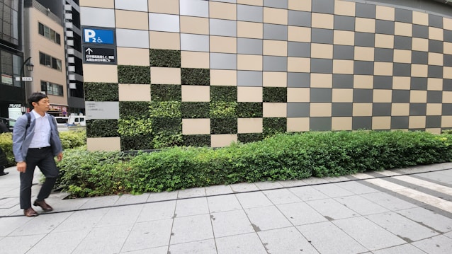 A man in a blue suit walks on a sidewalk beside a building facade made up of alternating beige and green checkered tiles. The lower part of the facade has green bushes. There is a blue parking sign with a wheelchair icon pointing to the left.