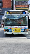 A public bus with a white, blue, and red color scheme is parked on a street. The front of the bus features a yellow sign with text and a cartoon character. Various signs, including one for wheelchair accessibility, are displayed on the bus. The street is in an urban area with buildings and shop signs in the background.