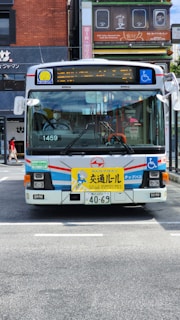 A public bus with a white, blue, and red color scheme is parked on a street. The front of the bus features a yellow sign with text and a cartoon character. Various signs, including one for wheelchair accessibility, are displayed on the bus. The street is in an urban area with buildings and shop signs in the background.