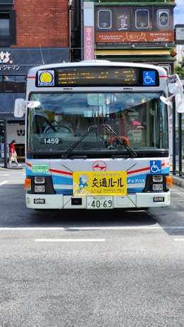A public bus with a white, blue, and red color scheme is parked on a street. The front of the bus features a yellow sign with text and a cartoon character. Various signs, including one for wheelchair accessibility, are displayed on the bus. The street is in an urban area with buildings and shop signs in the background.