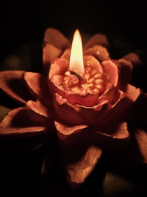 A single candle is lit, shaped like a blooming flower with intricate petals. The flame casts a warm, soft glow on the textured surface, creating a serene and intimate atmosphere.