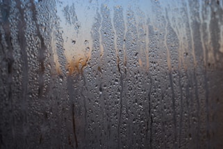 Condensation on a window creates intricate patterns with small droplets and streaks of water. The background appears blurred, with hints of warm colors possibly depicting a sunset or sunrise.