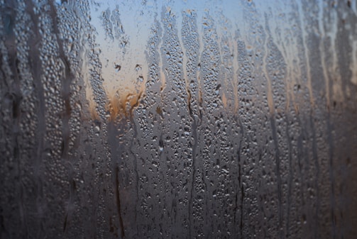 Condensation on a window creates intricate patterns with small droplets and streaks of water. The background appears blurred, with hints of warm colors possibly depicting a sunset or sunrise.