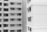 A black and white image showing the exterior of two multi-story residential buildings. One building is on the left and the other is on the right. Both buildings have numerous windows, some of which have open curtains or blinds. The architectural styles are modern with clean lines and rectangular shapes.