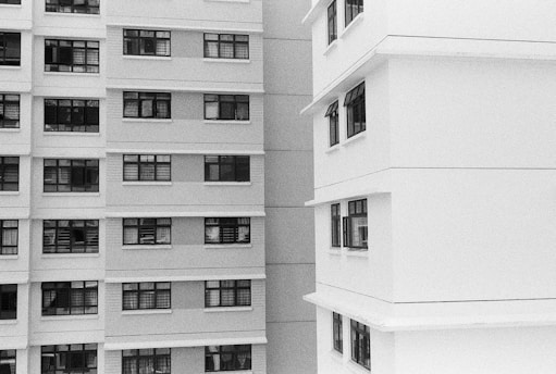 A black and white image showing the exterior of two multi-story residential buildings. One building is on the left and the other is on the right. Both buildings have numerous windows, some of which have open curtains or blinds. The architectural styles are modern with clean lines and rectangular shapes.