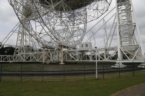 A large, intricate radio telescope structure dominates the scene, built from white metal beams and latticework. It stands against a backdrop of overcast sky and lush greenery. A fence runs along the foreground, separating the structure from the grassy area nearby.