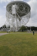 A large radio telescope with a circular dish supported by a complex network of white steel trusses dominates the scene. It stands amidst a green grassy field, with a few people visible in the distance. The sky is partly cloudy, and trees line the horizon.
