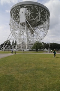 A large radio telescope with a circular dish supported by a complex network of white steel trusses dominates the scene. It stands amidst a green grassy field, with a few people visible in the distance. The sky is partly cloudy, and trees line the horizon.