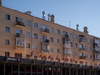 A multi-story residential building with balconies and a flat rooftop. Below the residential floors, there is a ground-level commercial area with signs in Cyrillic script. The building exterior has a beige color and several chimneys are visible on the roof.
