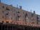 A multi-story residential building with balconies and a flat rooftop. Below the residential floors, there is a ground-level commercial area with signs in Cyrillic script. The building exterior has a beige color and several chimneys are visible on the roof.