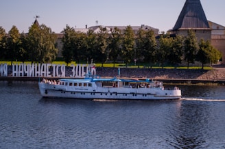 A white passenger boat is cruising on a calm river. In the background, there is a row of lush green trees and large letters forming a text sign along the riverside with an extensive wall of historic fortifications. The scene is bathed in pleasant daylight.