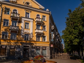 A multi-story building with a yellow facade, white balconies, and ornate architectural details. It faces a street lined with trees and features a sign with Cyrillic text. Flower beds adorn the front of the building, adding color to the scene under a clear blue sky.