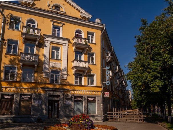 A multi-story building with a yellow facade, white balconies, and ornate architectural details. It faces a street lined with trees and features a sign with Cyrillic text. Flower beds adorn the front of the building, adding color to the scene under a clear blue sky.