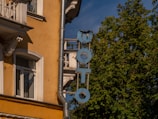 A vintage building with a mustard-yellow facade featuring ornate white window frames and decorative architectural elements. A vertical sign with the word 'PHOTO' in weathered blue letters is attached to the wall. Lush green trees and a clear blue sky are visible in the background.