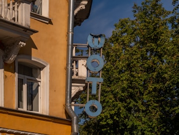 A vintage building with a mustard-yellow facade featuring ornate white window frames and decorative architectural elements. A vertical sign with the word 'PHOTO' in weathered blue letters is attached to the wall. Lush green trees and a clear blue sky are visible in the background.