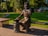 A bronze statue of a bearded blacksmith seated on a wooden bench, holding a shoe in one hand and a hammer resting on an anvil. The statue is situated in a park with green grass, trees, and colorful flowers in the background. A playground with various equipment can also be seen in the distance.