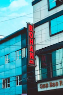 A street view of a modern building featuring a vertical red sign with the letters 'ROHAN ACC.' The building is composed of blue and gray panels with multiple windows. Power lines crisscross the scene, and part of a sign with 'GEE ESS F' is visible at the bottom.