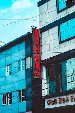 A street view of a modern building featuring a vertical red sign with the letters 'ROHAN ACC.' The building is composed of blue and gray panels with multiple windows. Power lines crisscross the scene, and part of a sign with 'GEE ESS F' is visible at the bottom.
