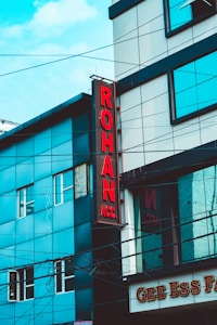 A street view of a modern building featuring a vertical red sign with the letters 'ROHAN ACC.' The building is composed of blue and gray panels with multiple windows. Power lines crisscross the scene, and part of a sign with 'GEE ESS F' is visible at the bottom.