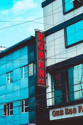 A street view of a modern building featuring a vertical red sign with the letters 'ROHAN ACC.' The building is composed of blue and gray panels with multiple windows. Power lines crisscross the scene, and part of a sign with 'GEE ESS F' is visible at the bottom.