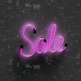 Pink neon light word 'Sale' is displayed against a textured dark background, with glow reflecting off the surface.