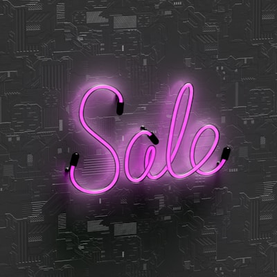 Pink neon light word 'Sale' is displayed against a textured dark background, with glow reflecting off the surface.