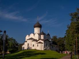 A historic Orthodox church with distinctive onion domes stands on a grassy hill. The building's architecture includes white walls and intricate designs, surrounded by lush green trees. A clear blue sky forms the backdrop, and a group of people is gathered near the church.