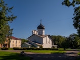 A white, historic Orthodox church with a dark domed roof is situated in a lush, green park setting. There are trees surrounding the area, and a few other buildings in the background. The sky is clear and blue, indicating a sunny day.