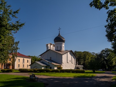 A white, historic Orthodox church with a dark domed roof is situated in a lush, green park setting. There are trees surrounding the area, and a few other buildings in the background. The sky is clear and blue, indicating a sunny day.