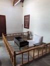 A simple, traditional room with minimal furniture, featuring a low wooden bed and desk, a spinning wheel, and a wall poster with text and a circular design. The space is enclosed by a wooden railing and has a closed wooden door on one side.